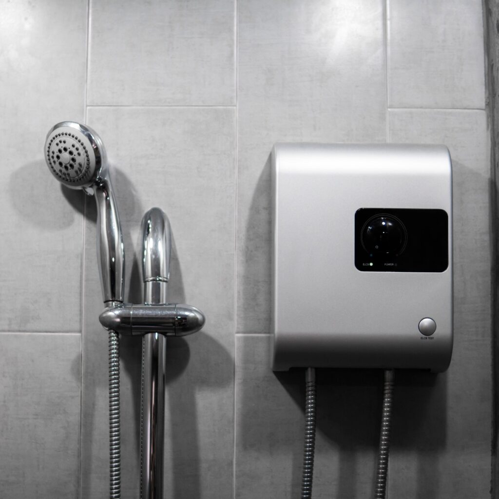 Instant tankless electric water heater installed on grey tile wall with input and output pipe outlet and elcb safety breaker system and silver shower.
