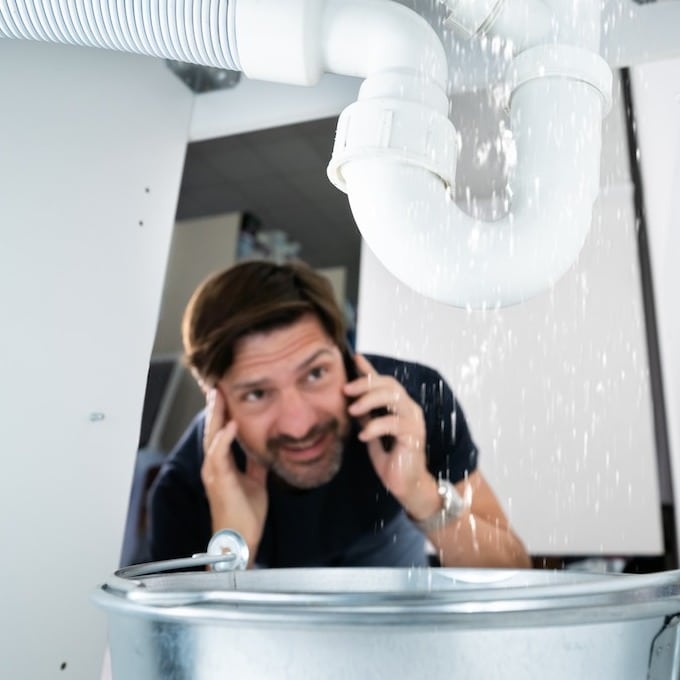 A male homeowner is looking at a PVC pipe under his sink that is dripping into a bucket while hold a phone to his ear calling a plumber.